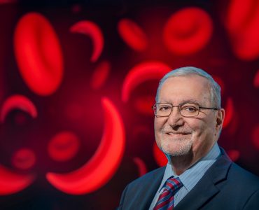 Man smiles at camera with image of red blood cells and sickle cells behind him.