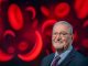 Man smiles at camera with image of red blood cells and sickle cells behind him.